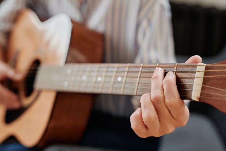 Como aprender a tocar violão do zero, mesmo sem professor Close das mãos de uma pessoa pressionando as cordas no braço de um violão acústico durante a prática