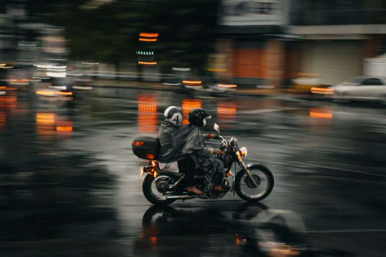 Delivery em dias de chuva sobe até 7,2% e não cobre a queda de 34,4% no salão Dois motociclistas de capa de chuva atravessam em movimento uma avenida molhada, com luzes alaranjadas refletidas no asfalto